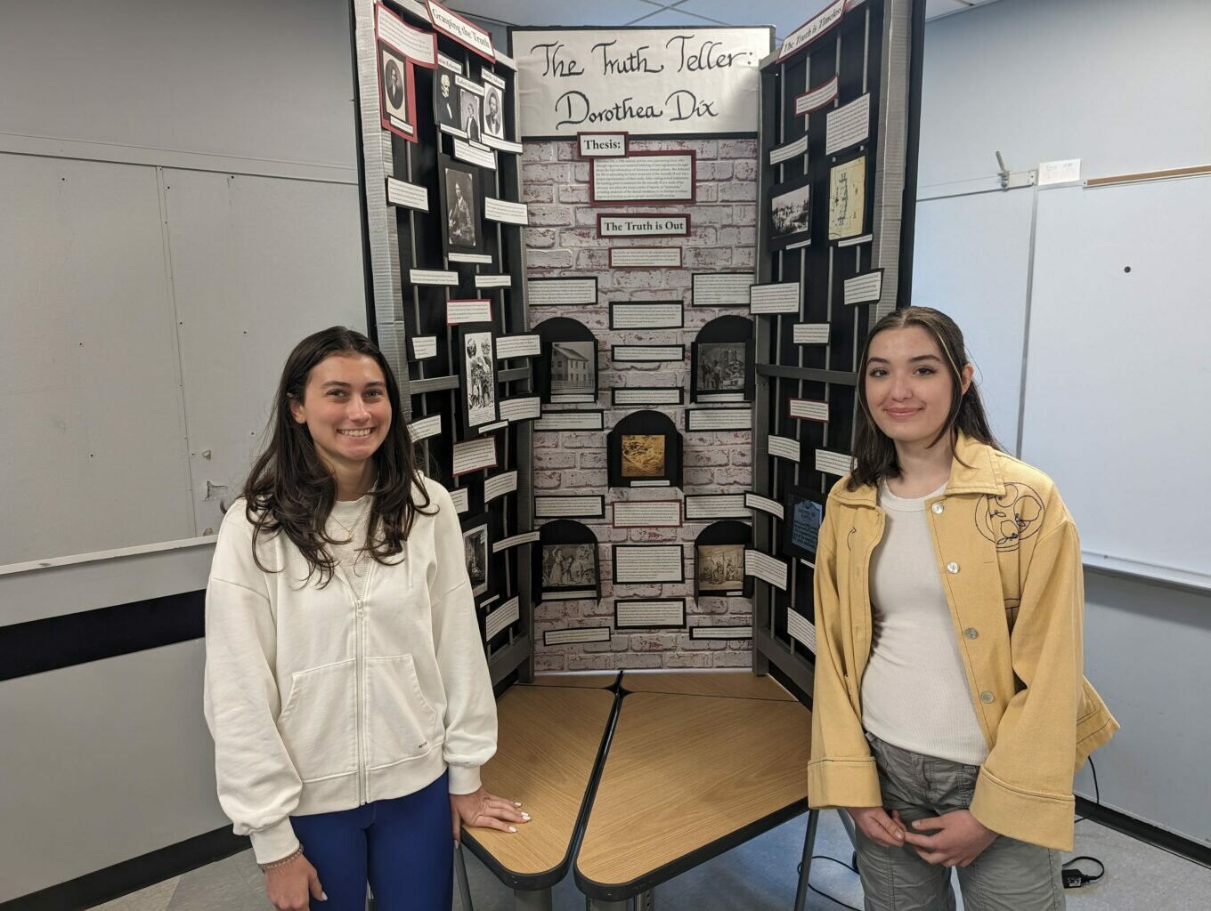 Two female students stand smiling in a classroom next to their project display titled "The Truth Teller: Dorothea Dix." The display is a three-panel board with a brick-patterned center, featuring various historical photos, a thesis statement, and informational text boxes arranged to resemble window panes or cells. The student on the left wears a white hoodie and blue pants, while the student on the right wears a yellow jacket over a white shirt.