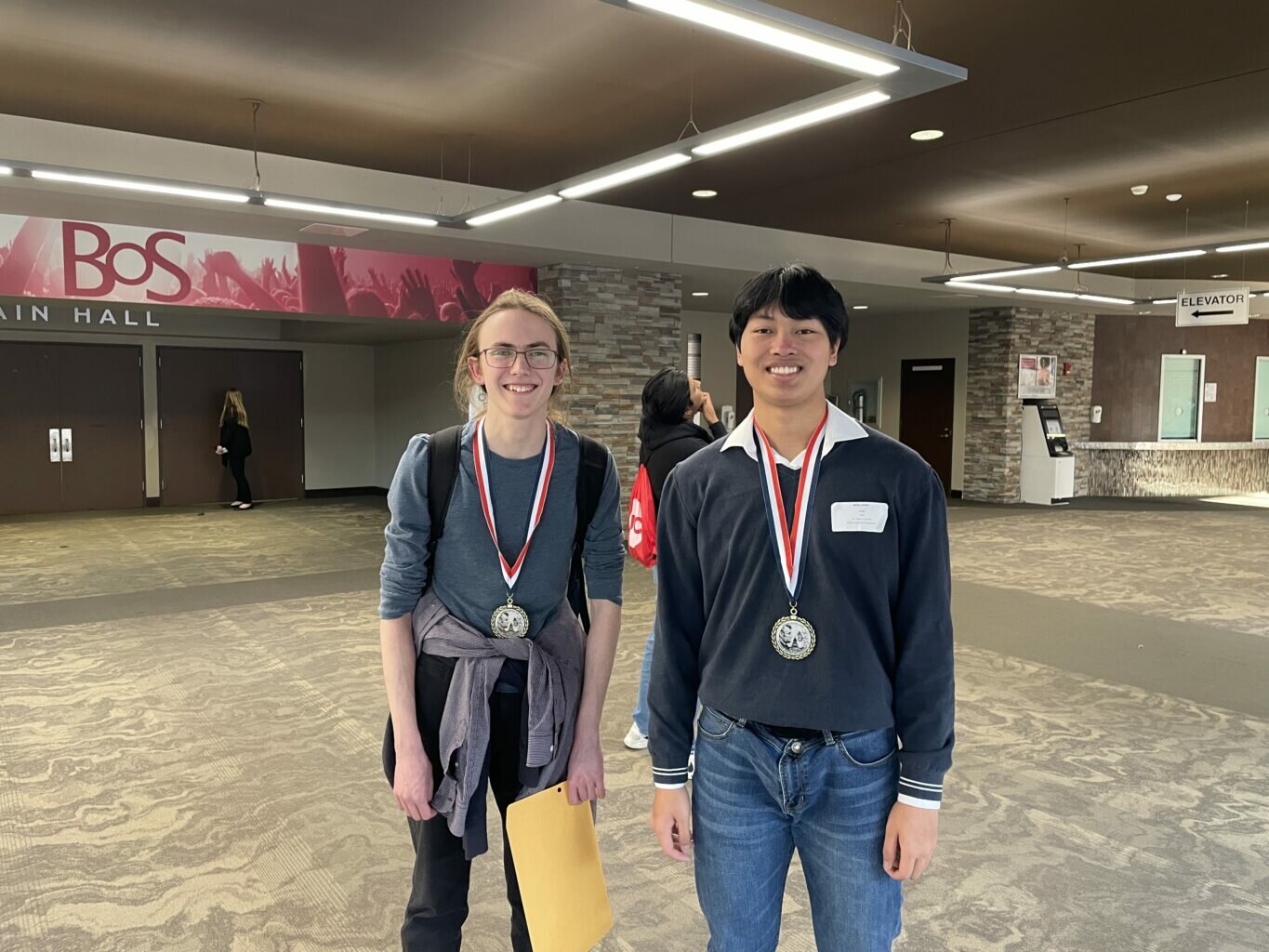 Two smiling male students stand in a spacious lobby wearing large silver medals with red, white, and blue ribbons. The student on the left wears glasses, a blue long-sleeve shirt, and has a jacket tied around his waist; the student on the right wears a dark sweater over a collared shirt and blue jeans. In the background, a sign reads "BOS Main Hall."