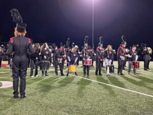 Members of the Unified drumline perform on a high school football field at night, wearing black and red uniforms and playing various percussion instruments under stadium lights.