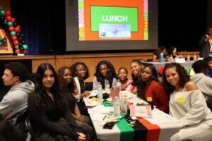 Students sit at a table decorated with Pan-African flag colors during a school lunch event featuring a presentation on Jamaican cuisine.