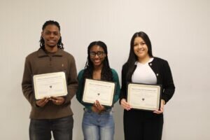 Three people stand side by side, smiling and holding certificates in front of a plain white wall. They appear to have received awards or recognition.
