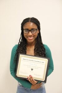 A young woman with long braided hair and glasses smiles while holding a framed certificate in front of a plain background.