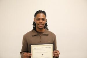 A smiling person with long braided hair, wearing a brown zip-up sweater, holds up a framed certificate against a plain light background.