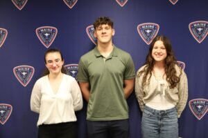 Three teenagers stand side by side in front of a blue backdrop with repeating District 207 logos, smiling at the camera. 