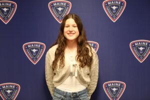 A young woman with long brown hair, wearing a white top, gray knit cardigan, and blue jeans, stands smiling in front of a blue backdrop with repeating District 207 logos