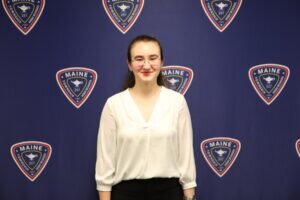 A person with long brown hair, glasses, and a white blouse stands smiling in front of a blue backdrop with repeating District 207 logos