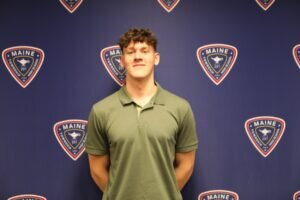A young man with curly brown hair wearing a green polo shirt stands smiling in front of a blue backdrop with multiple District 207 logos printed on it.