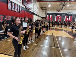 A high school band plays saxophones in a gym, standing in a line on the basketball court. Behind them are bleachers and a red-and-black wall with HAWKS written in large white letters.