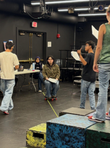 Several teens rehearse in a black box theater. One sits on a skateboard holding a script, others stand or sit nearby reading. Stage blocks are in the foreground, and a table with two people is in the background.