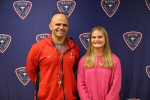 A man in a red zip-up jacket and a woman in a pink sweater stand together in front of a blue backdrop featuring the Maine Township High School logo.