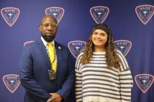 A man in a suit and yellow tie stands next to a woman in a striped sweater in front of a blue backdrop with Maine Township High School logos.