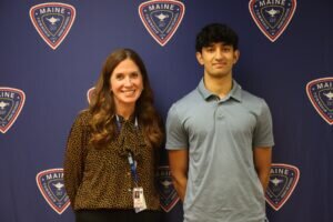 A female staff member and a male student stand in front of a blue backdrop with the Maine Township High School District 207 logo.