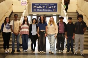 A teacher and eight students stand in a row in front of a Welcome to Maine East High School sign at the top of a staircase.