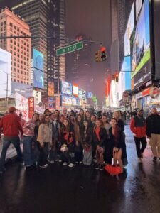 A large group of people stand together on a wet street in Times Square at night under illuminated billboards and a W 47 St street sign.