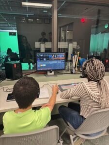 Two students wearing headsets work at a computer station in a television studio control room, editing video on a monitor.