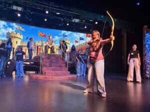 A student on a theater stage aims a bow and arrow toward the audience, with other cast members standing on a castle-themed set behind her.