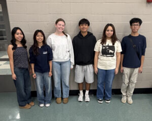 Six students standing in a row against a light-colored brick wall.
