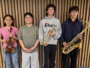 Four students standing in a row against a wooden slat wall; the first holds a violin, the second stands with hands clasped, the third holds a trumpet, and the fourth holds a saxophone.